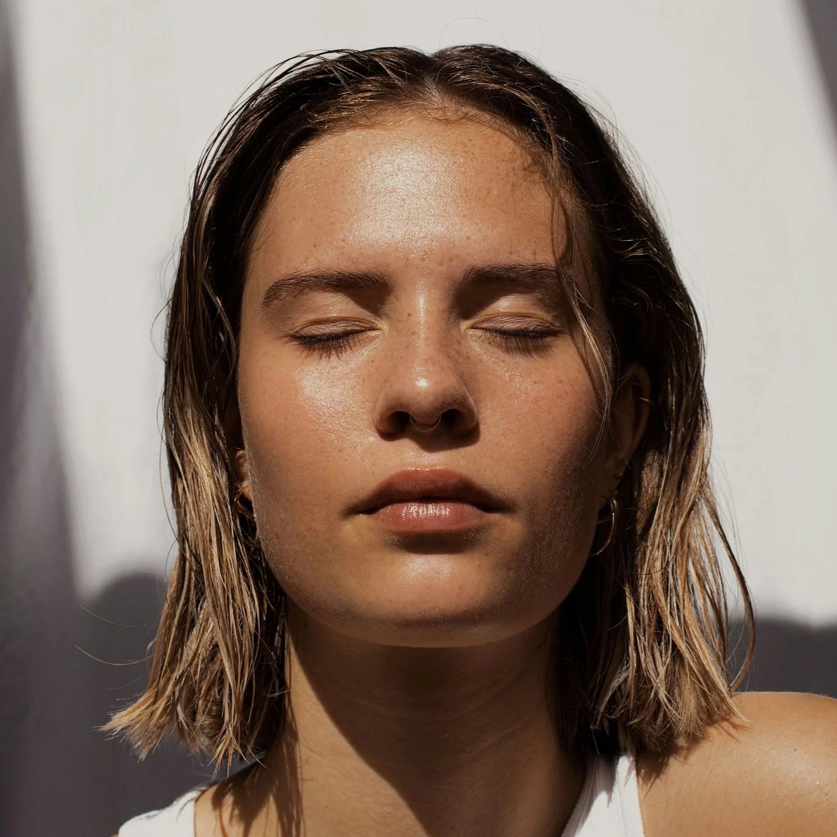 Close-up of a young woman with closed eyes, natural makeup, and wet hair, illuminated by sunlight.